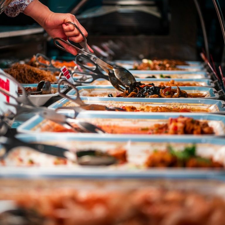 Buffet mit verschiedenen Gerichten, eine Hand nimmt Essen mit Zangen.